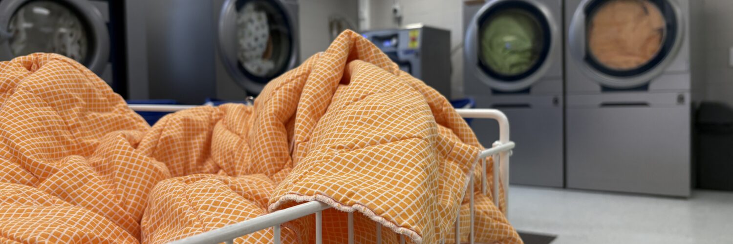 Kuvassa etualalla kori, jossa oranssi peitto ja taustalla näkyy neljä pyykinpesukonetta, jotka ovat täynnä pyykkiä.